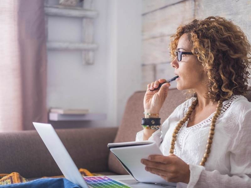 Photo of a woman sitting on a couch with a laptop on her lap. She is looking away from the camera biting a pen thoughfully